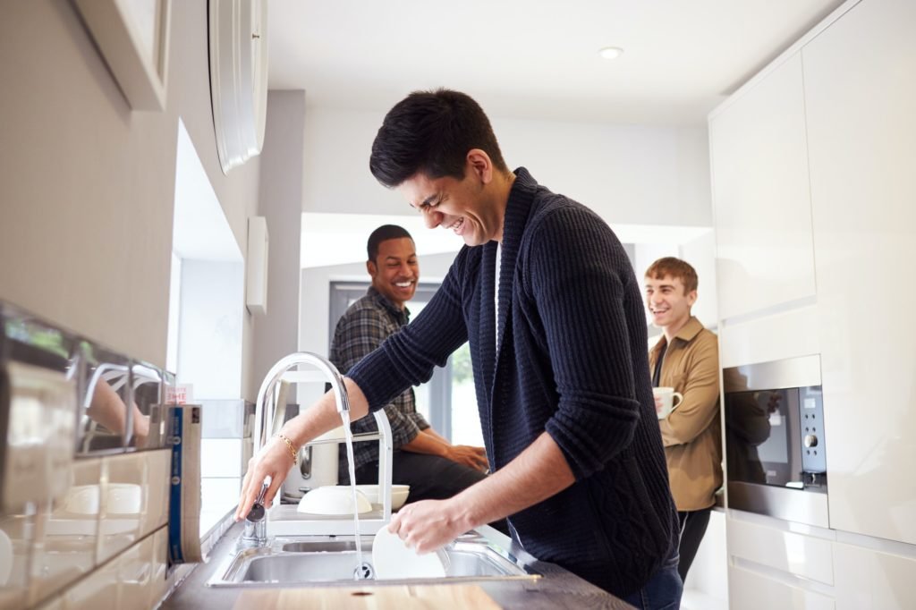 Group Of Male College Students In Shared House Kitchen Washing Up And Hanging Out Together