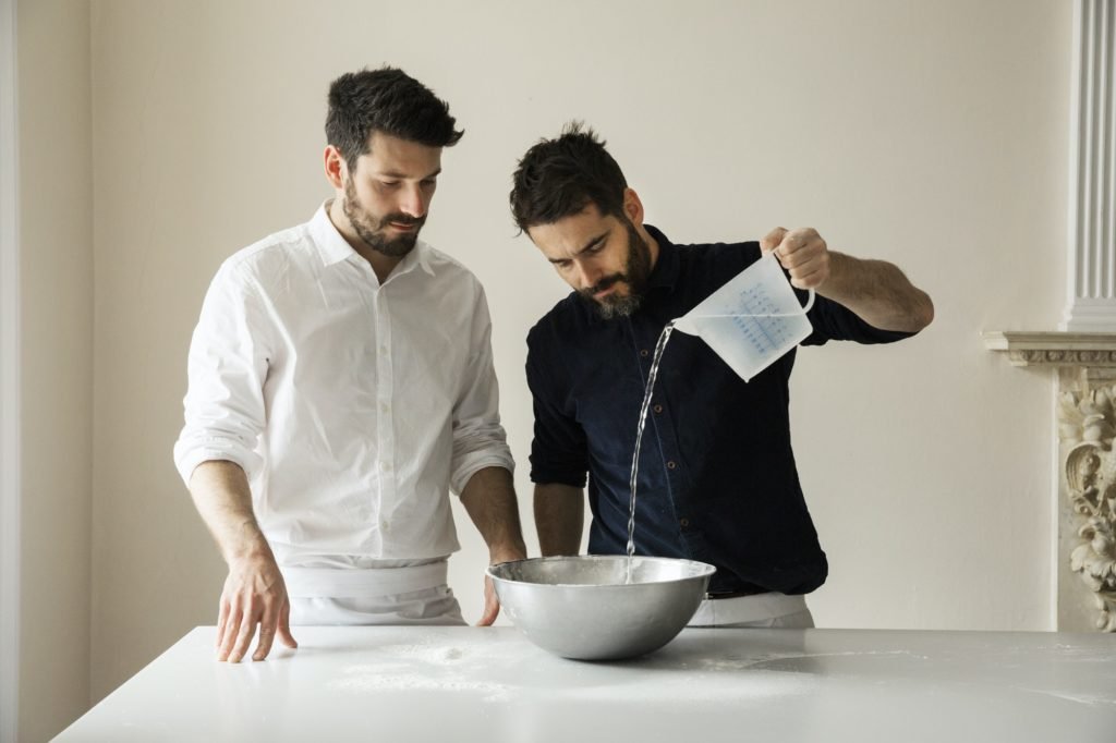 Two bakers preparing bread dough, pouring water from a measuring jug into a metal mixing bowl.