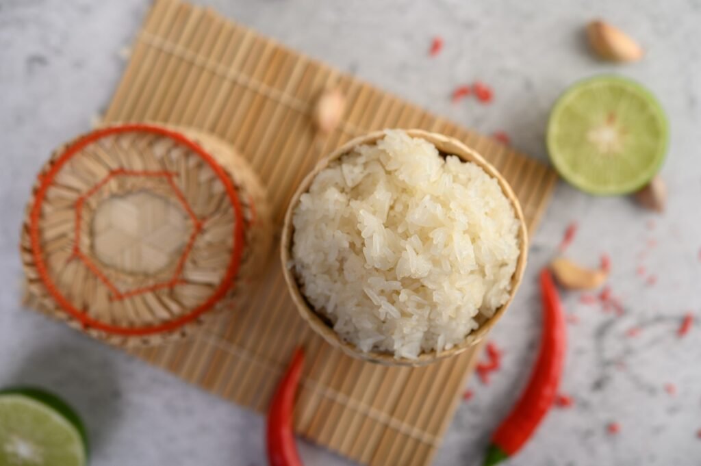 Thai sticky rice in a woven bamboo basket on a wooden panel with chilies, lime, and garlic.