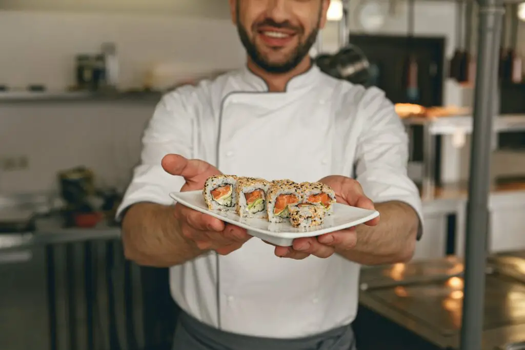 Handsome chef of japanies restaurant showing plate with sushi standing on kitchen