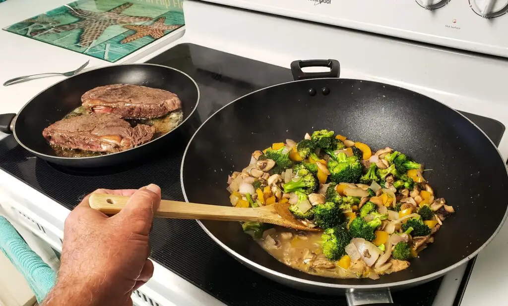 Man stirs vegetables Ina wok while steaks are seared on both sides for tasty dinner stovetop.