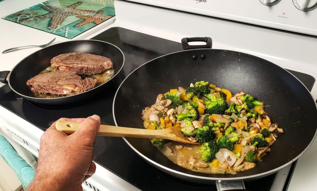 Man stirs vegetables Ina wok while steaks are seared on both sides for tasty dinner stovetop.