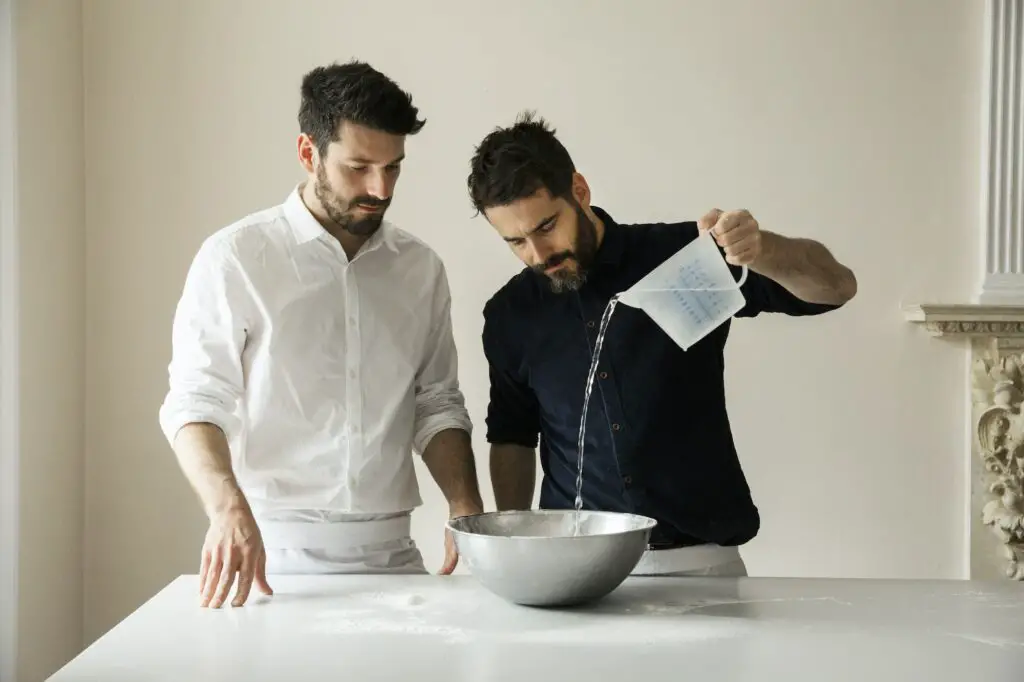 Two bakers standing at a table, preparing bread dough, pouring water from a measuring jug into a