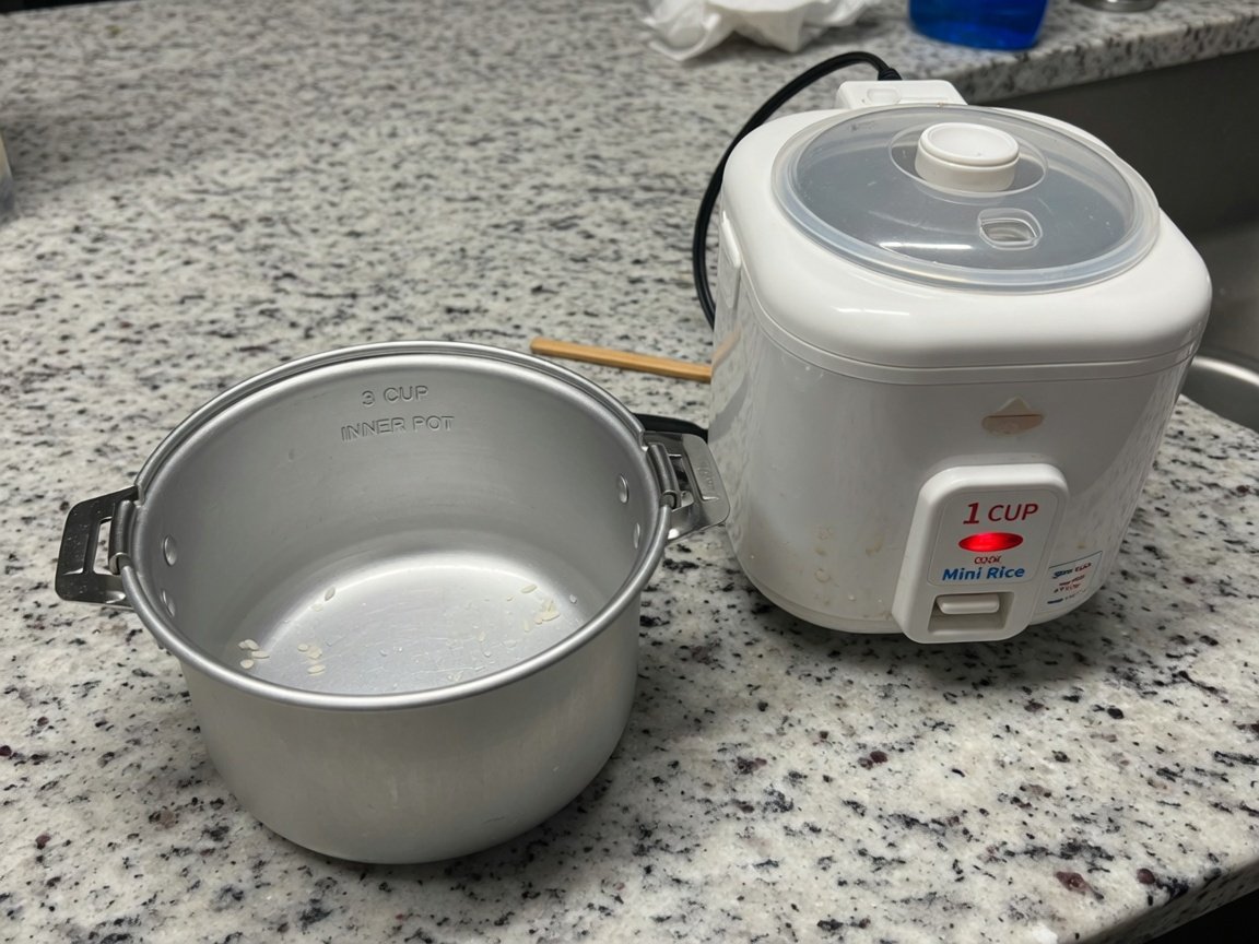 A small compact aluminum inner cooking pot photographed next to a mini single-serve rice cooker on a kitchen counter
